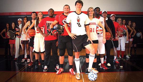 student athletes from multiple sports stand in a V-formation in the gym holding various sporting equipment